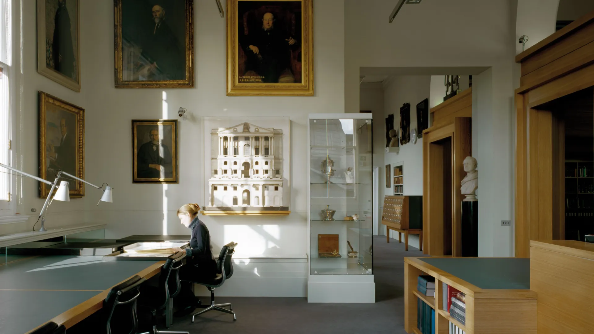 Lady working at a desk in the RIBA Reading Rooms at the V&A Museum with a lamp and large architecture models and paintings on the wall