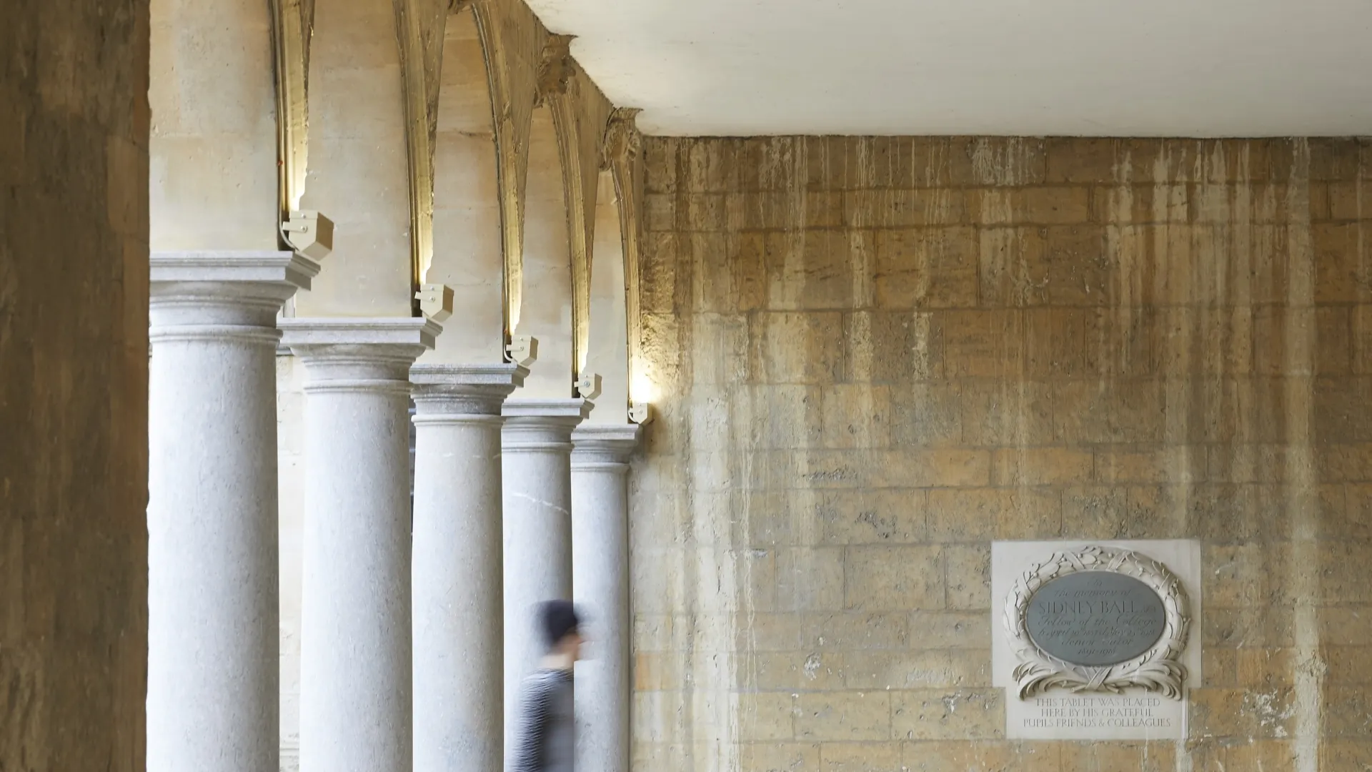 Person walking left to right in a stone colonnaded cloister.