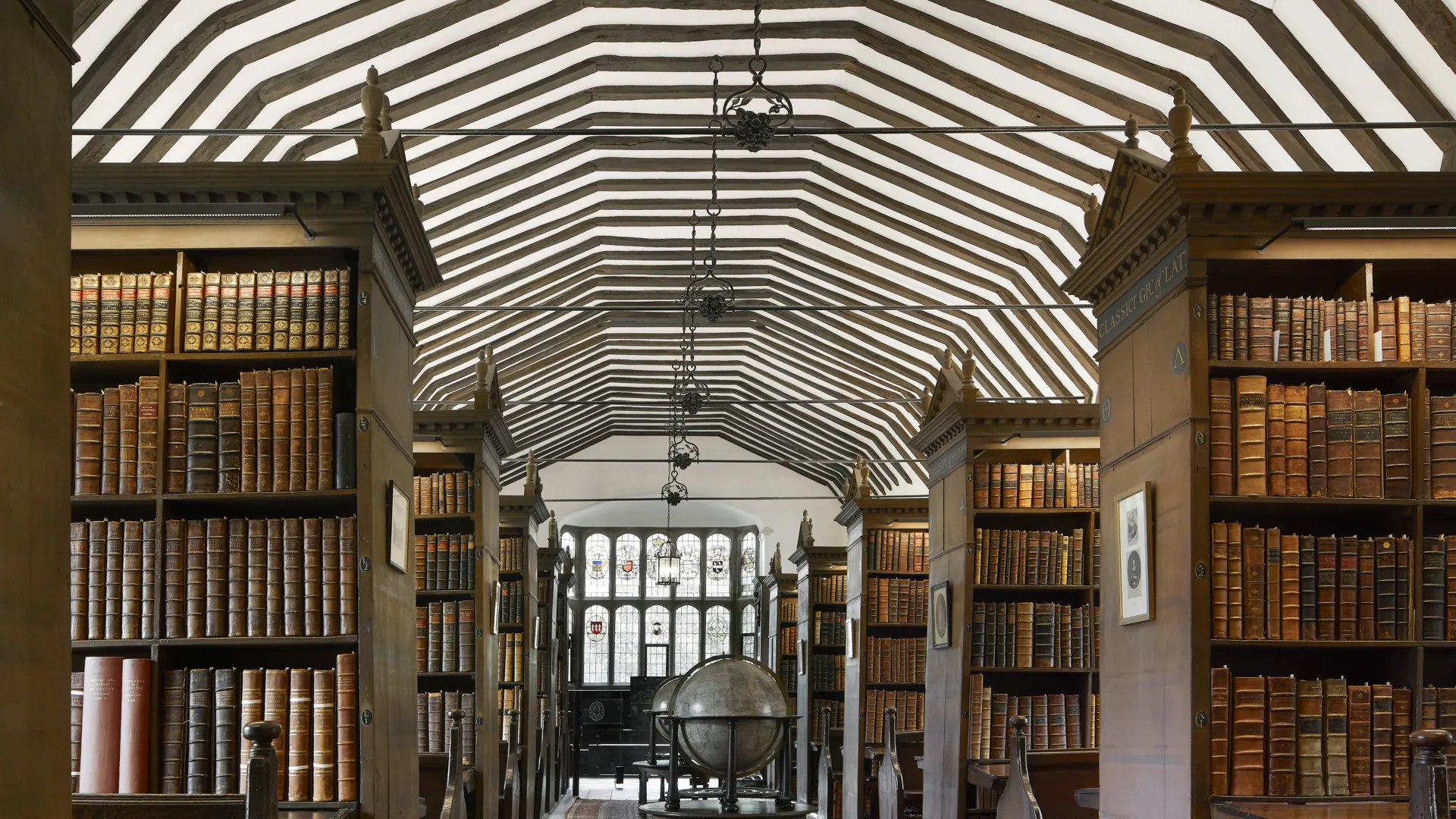 Historic library with bookshelves carpets and old globes beneath a wooden beamed ceiling.