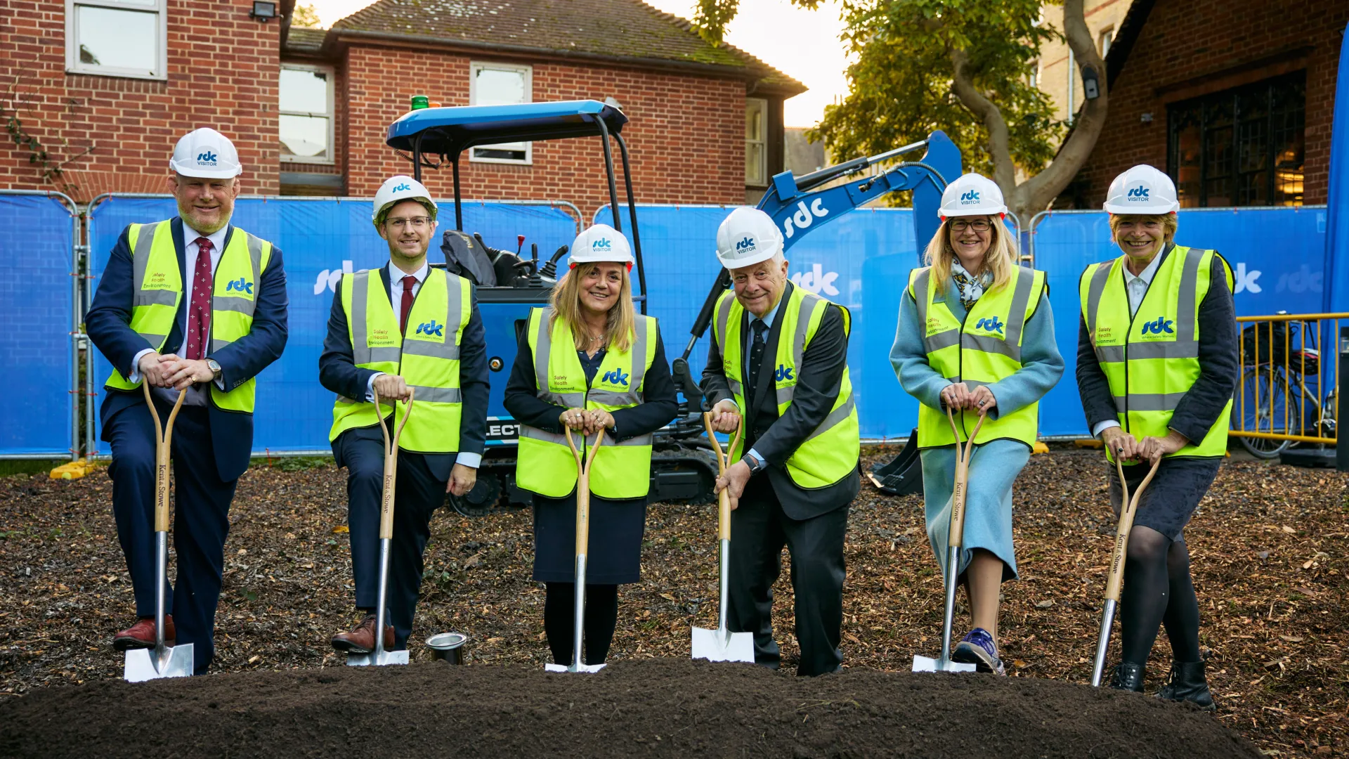 Six individuals in yellow safety vests stand on a mound of dirt with shovels in a groundbreaking ceremony.