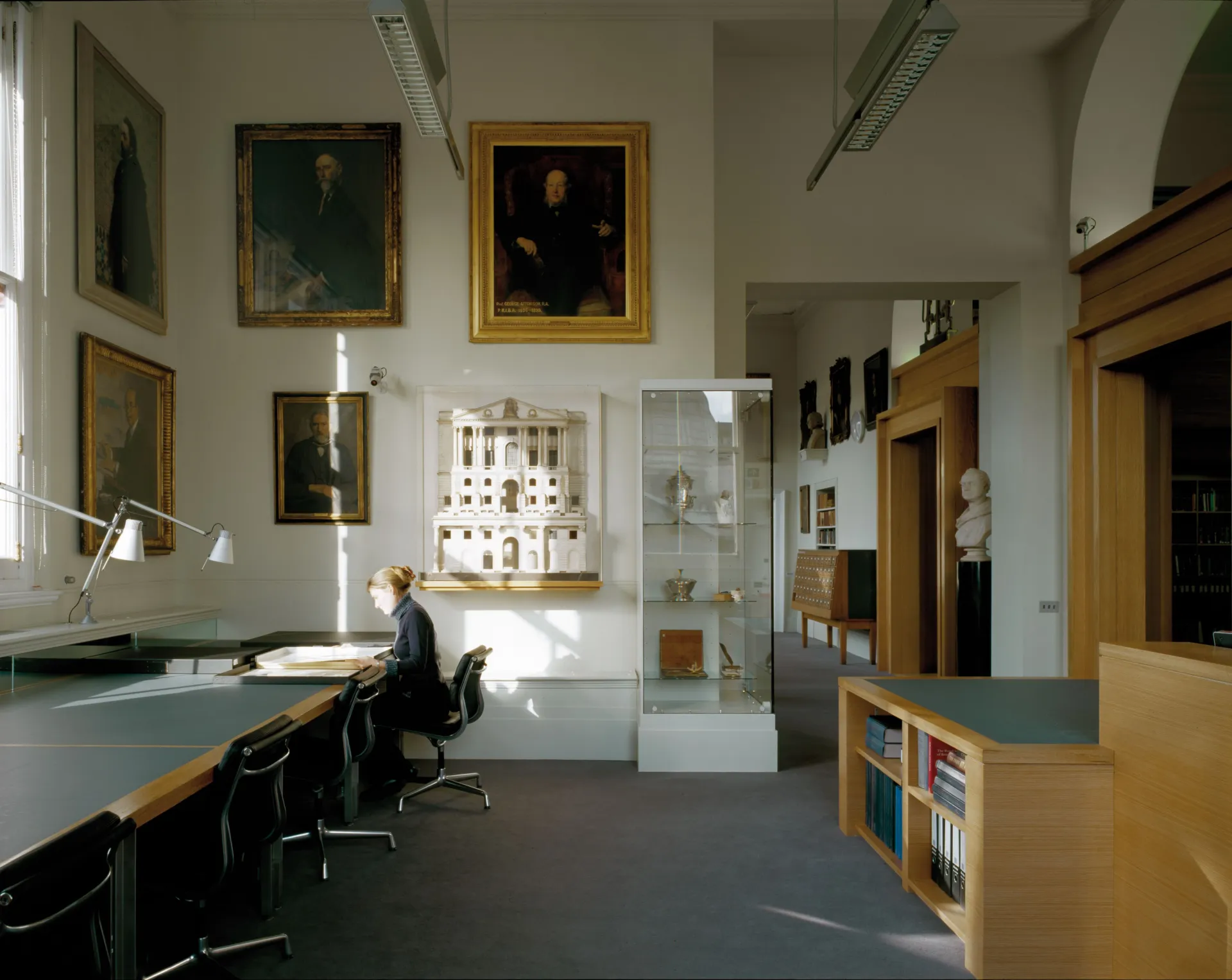 Lady working at a desk in the RIBA Reading Rooms at the V&A Museum with a lamp and large architecture models and paintings on the wall