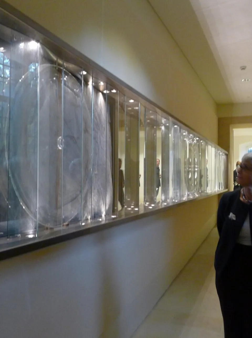 Two individuals stand at right facing left and looking at a glass art work installed in the wall.