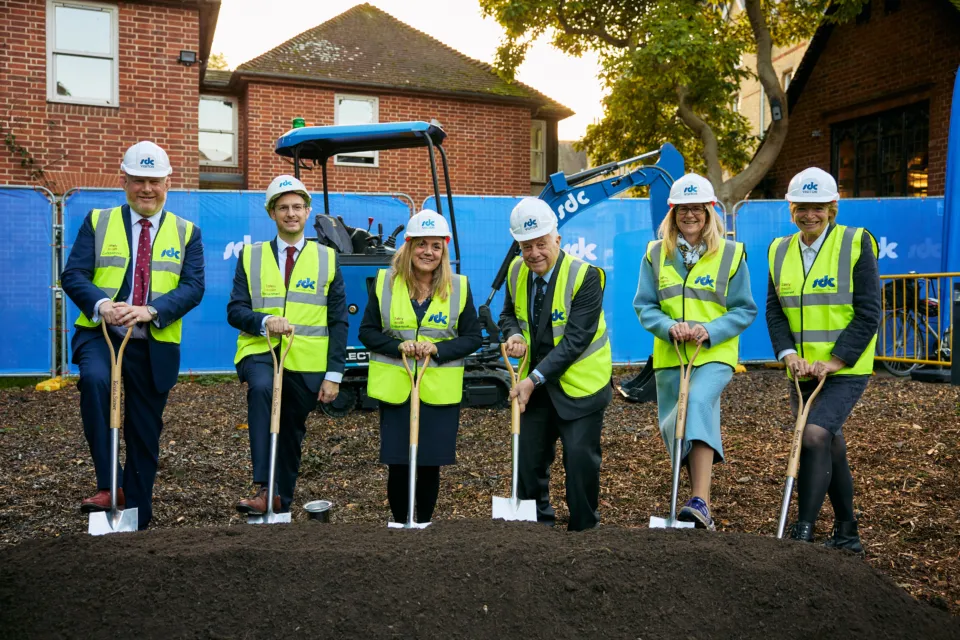 Six individuals stand in yellow protective vests on a mound of dirt with shovels in the soil.